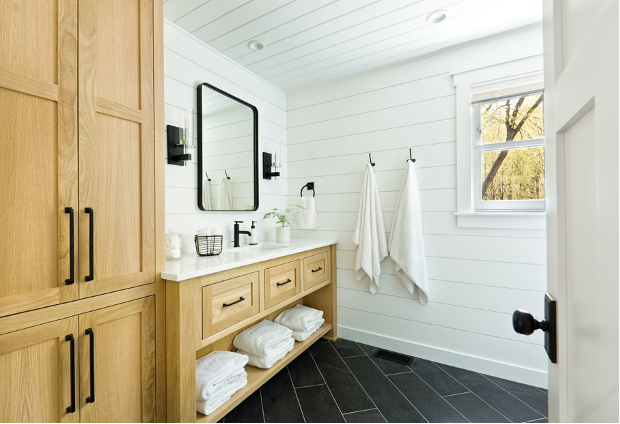 Bright bathroom with white shiplap walls, wood vanity, black mirror, and dark tile floor.