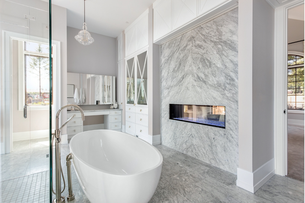 Bright modern bathroom with freestanding tub, marble fireplace, glass shower, and white cabinetry