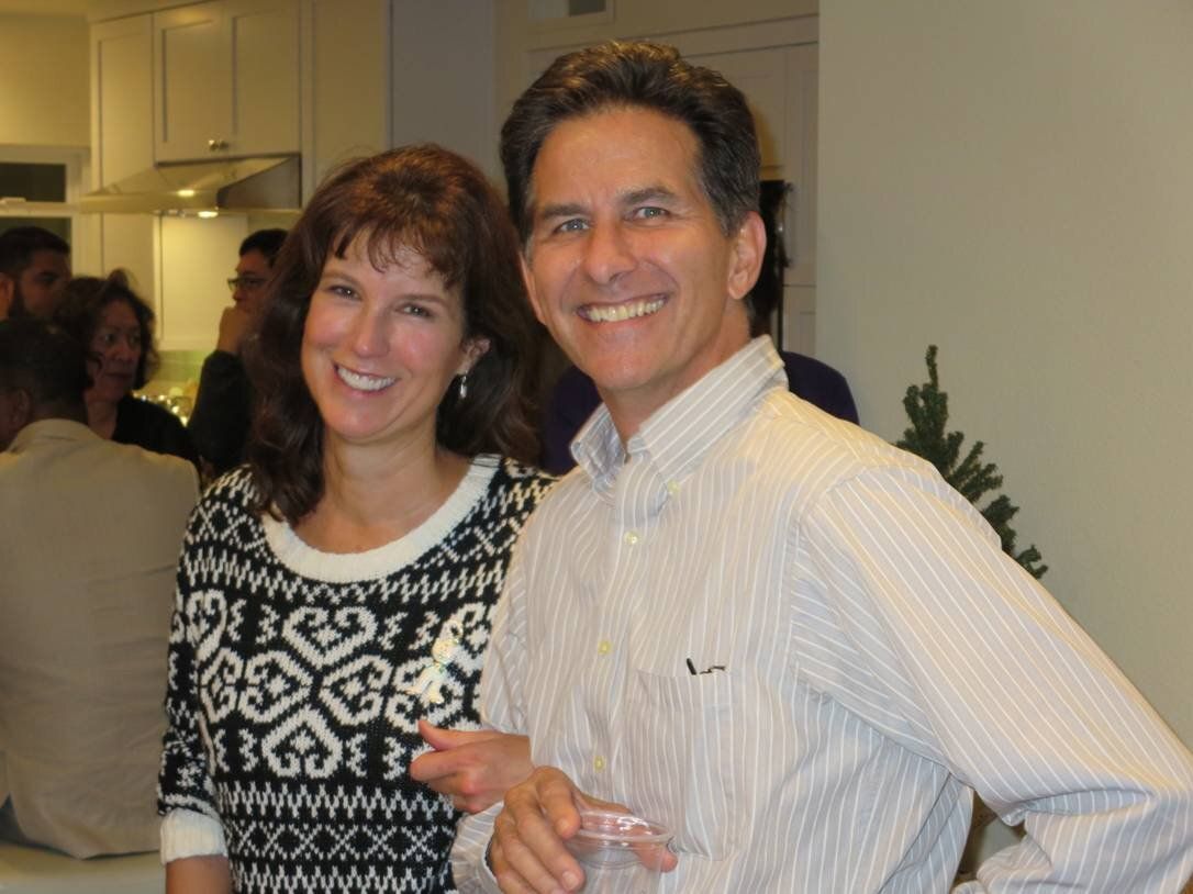 Two smiling people posing indoors at a social gathering, one holding a drink.