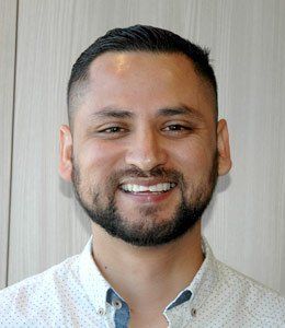 Smiling man with short dark hair and beard, wearing a white button-up shirt against a light wood background