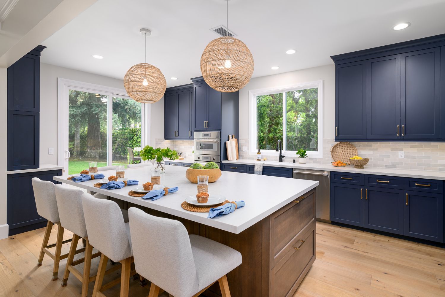 image of a large open floor plan kitchen with a large center island with bar height chairs, navy blue perimeter cabinets, medium brown stained island cabinetry, Omega Full Access cabinetry, white quartz countertops, light gray and white subway tile backsplash, stainless steel Thermador appliances, new hardwood floors French Oak, woven pendant light fixtures over island, Milgard Window, Milgard sliding back door, Agoura Hills, CA modern style Kitchen Remodel