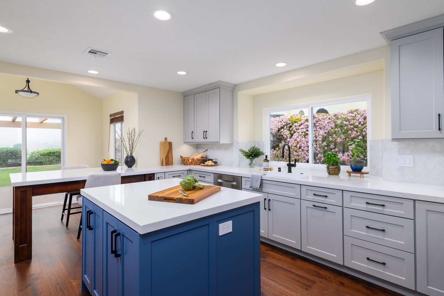 Bright kitchen with blue island, white cabinets, wood floors, and large windows overlooking greenery