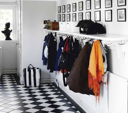 Hallway with black-and-white checkered floor, wall hooks with coats, framed photos, and a black cat on a shelf