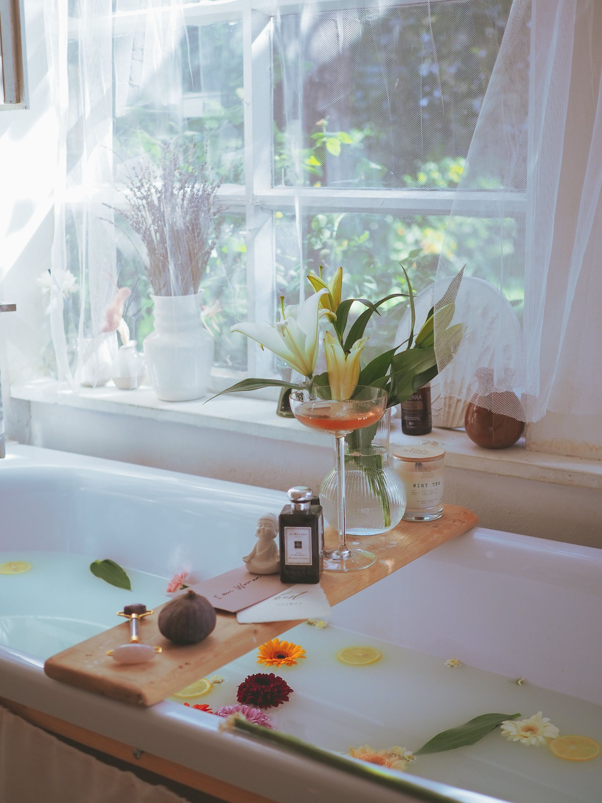 Bath window with flowers and candles on a tray beside a white tub, sunlight through lace curtains