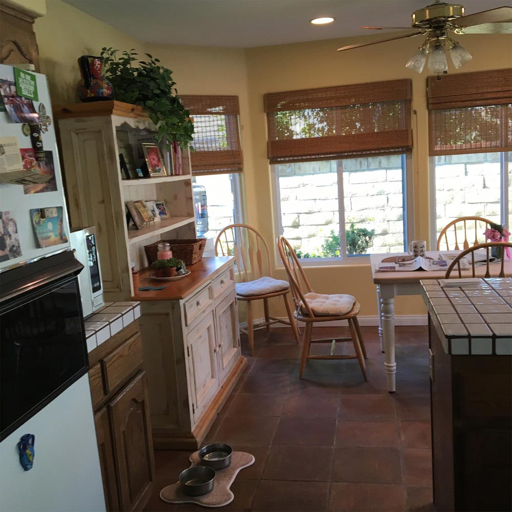 Bright kitchen with tiled floor, windows with blinds, wooden cabinets, and a small dining area by the wall.