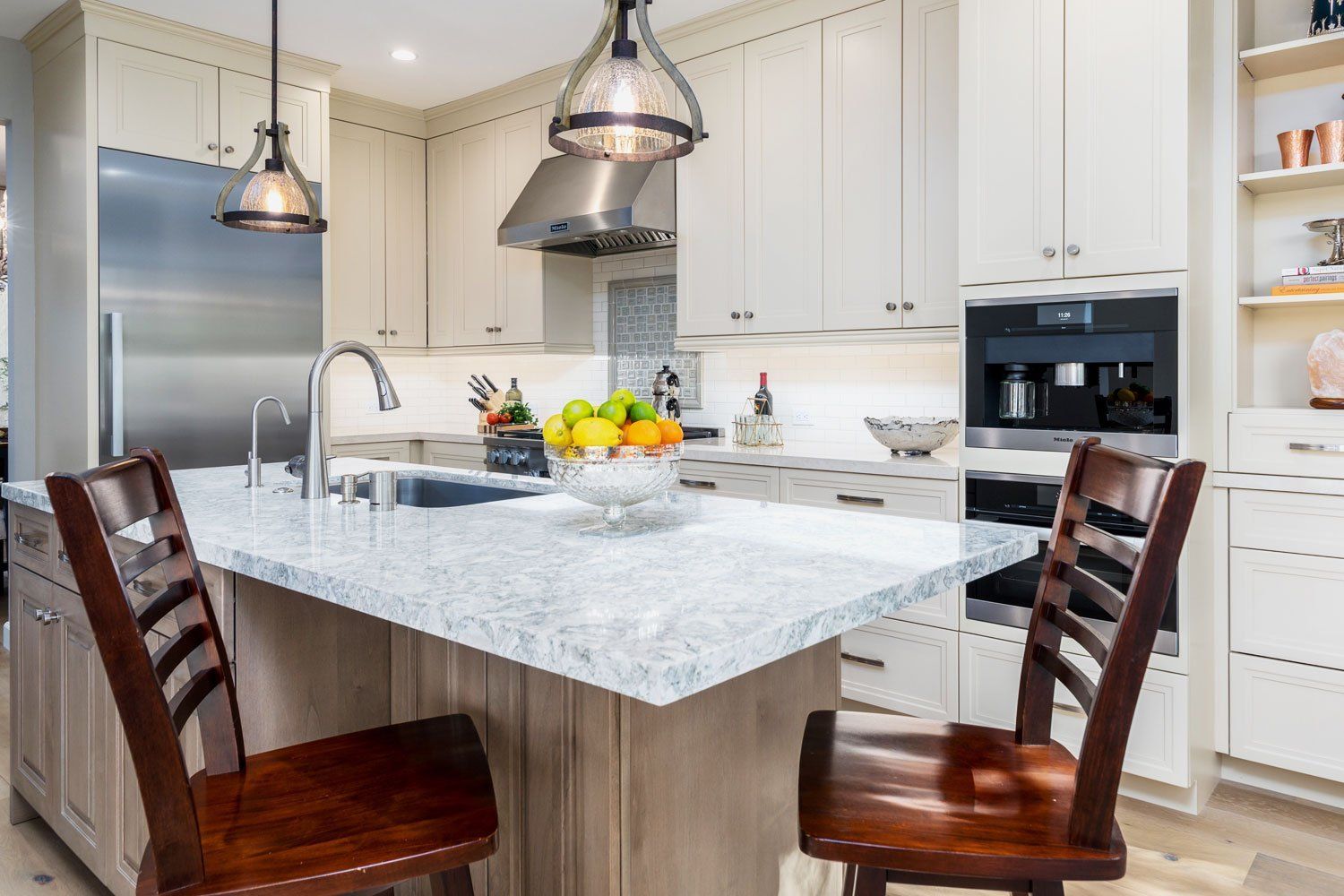 Bright modern kitchen with marble island, pendant lights, white cabinets, and two wooden stools