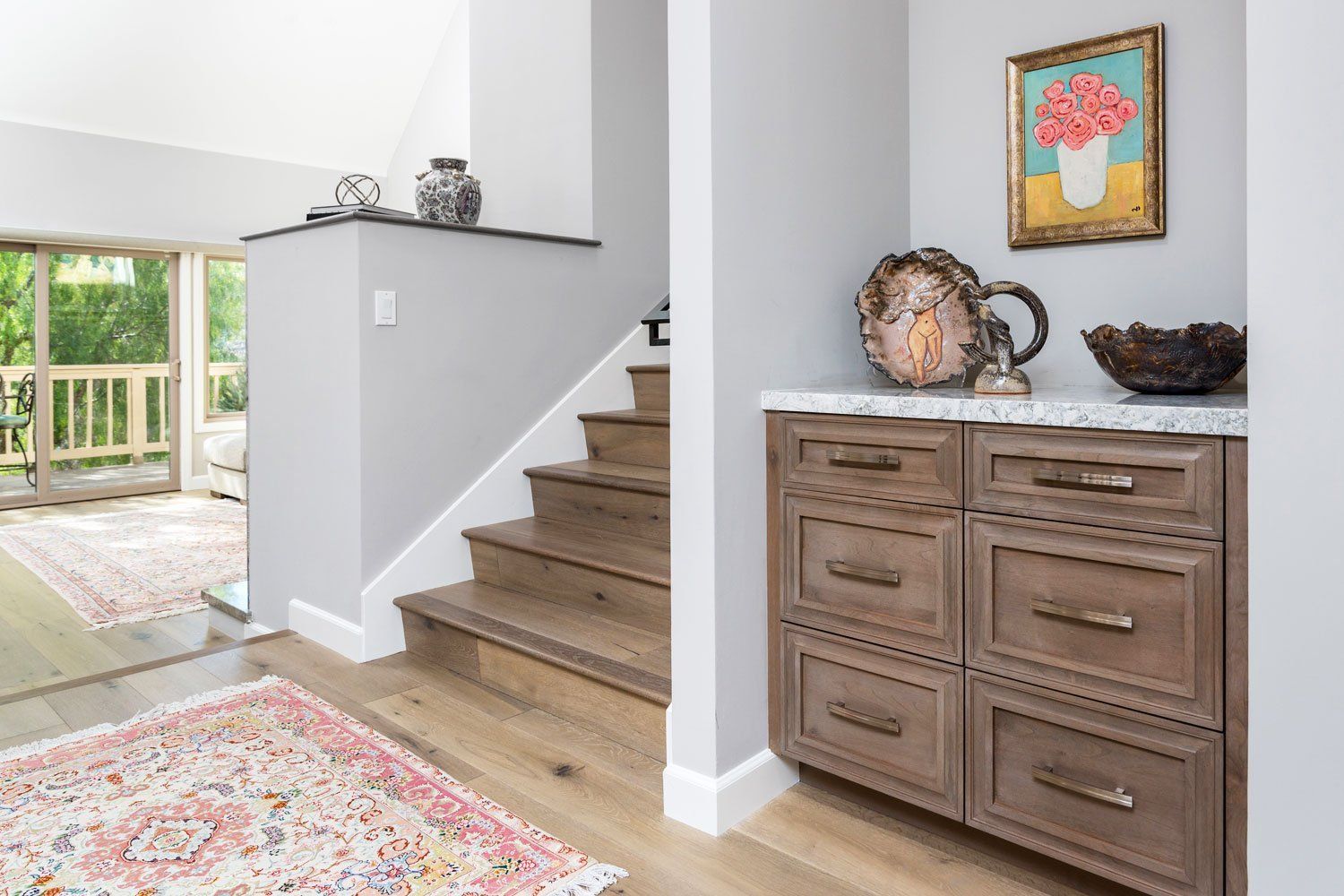 Bright entryway with wooden stairs, rustic cabinet, and rug beside a sunlit living area