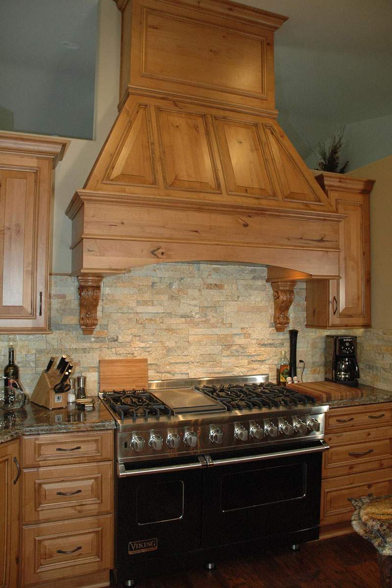 Rustic kitchen with stone backsplash, wood cabinets, and a black range with large wooden hood