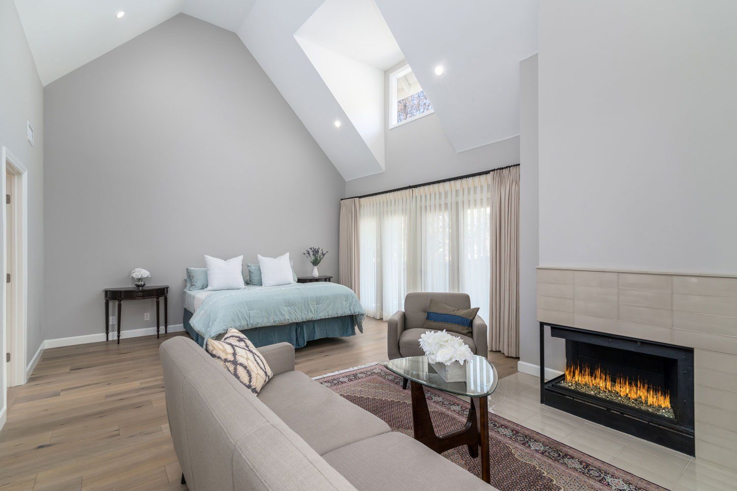 Bright modern living room with vaulted ceiling, fireplace, gray sofa, teal bed, and large windows
