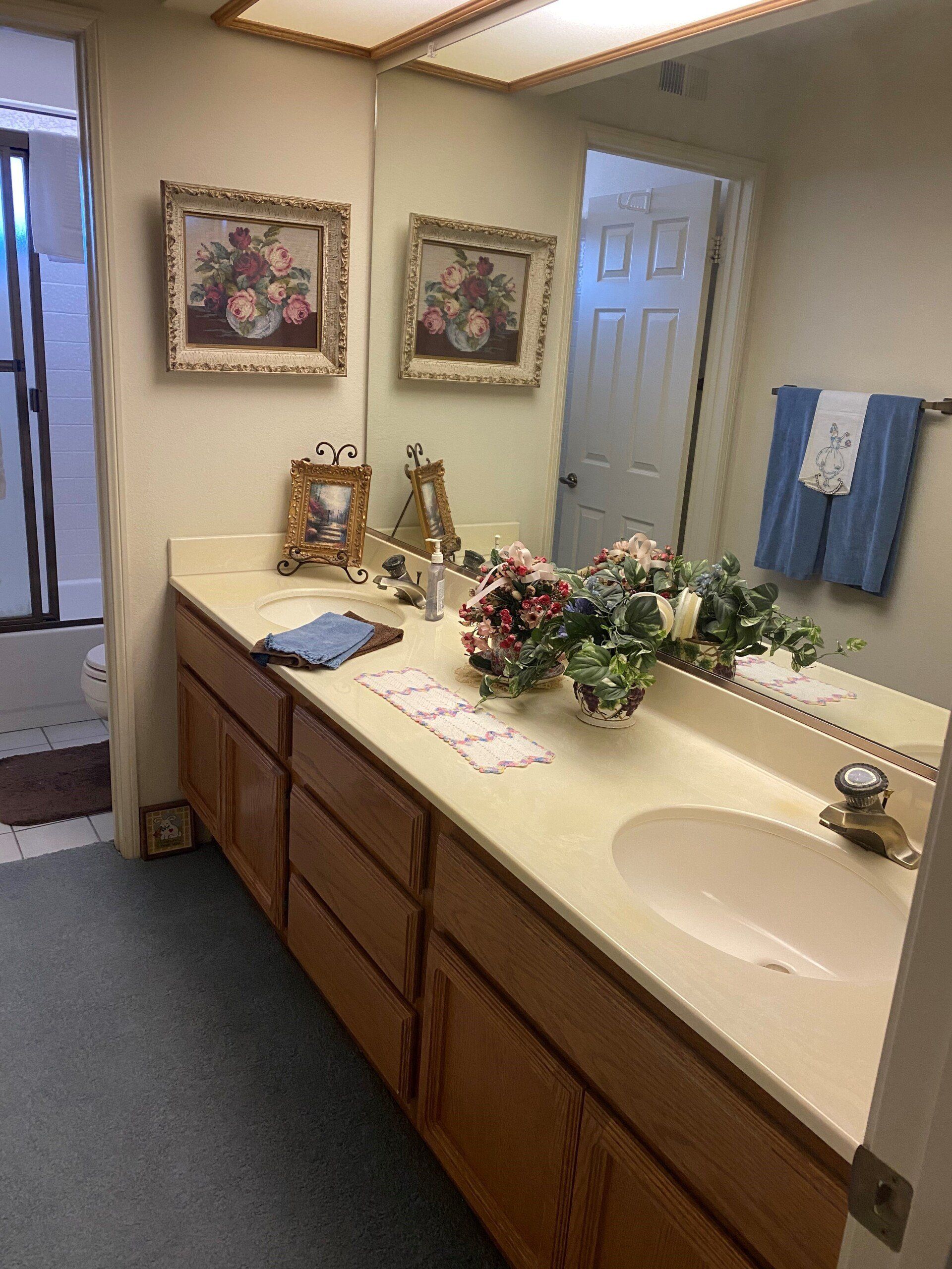 Bathroom vanity with double sinks, beige counters, flowers, mirror, and blue towels on the wall