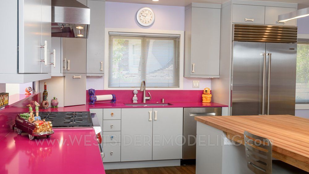 Modern kitchen with bright pink countertops, gray cabinets, stainless steel fridge, and a large window under a clock