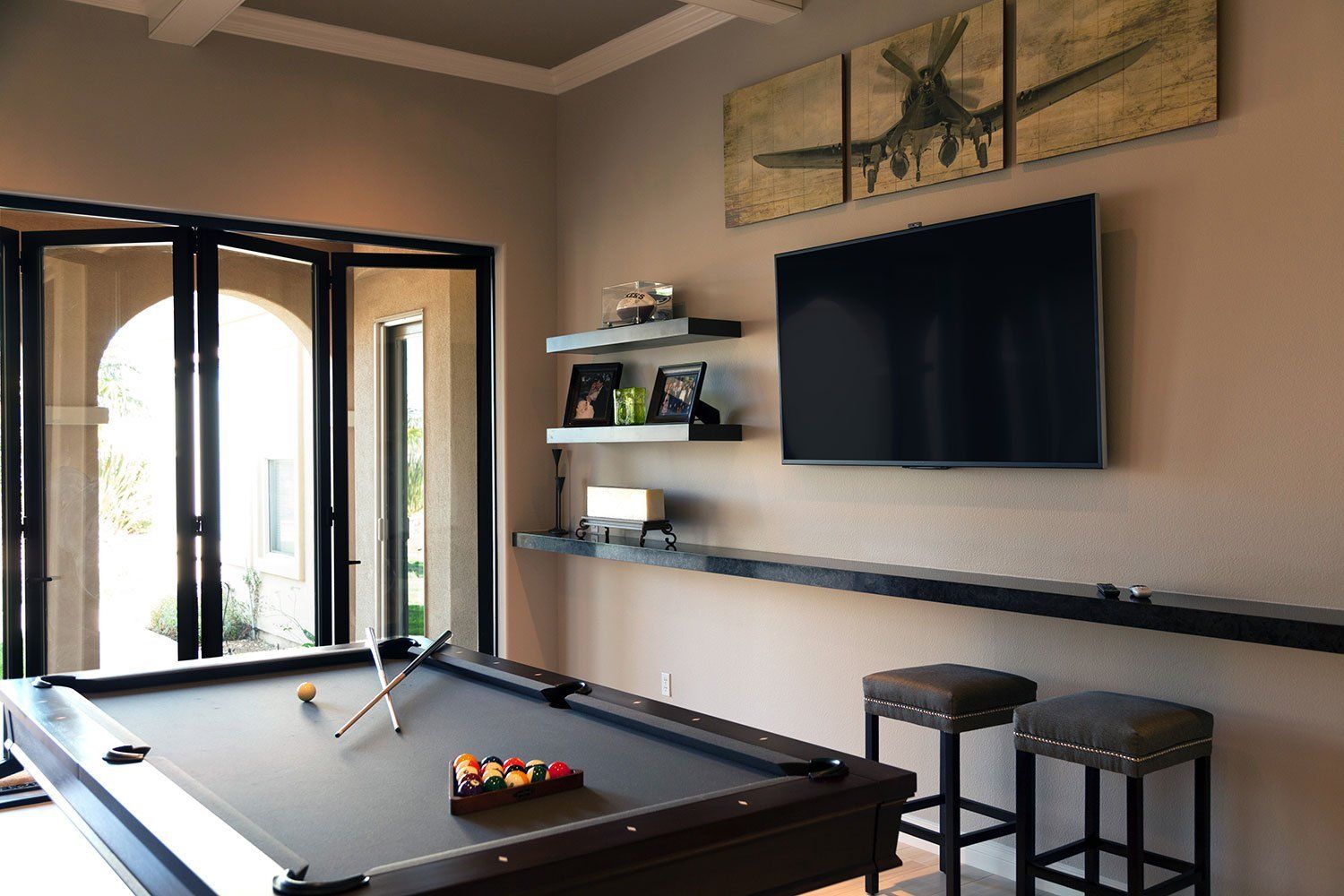 Bright game room with pool table, wall-mounted TV, bar shelves, stools, and open glass doors.