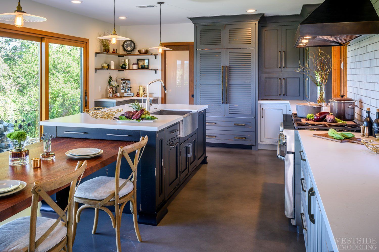 Modern kitchen with large island, blue-gray cabinets, wood table, and bright windows.