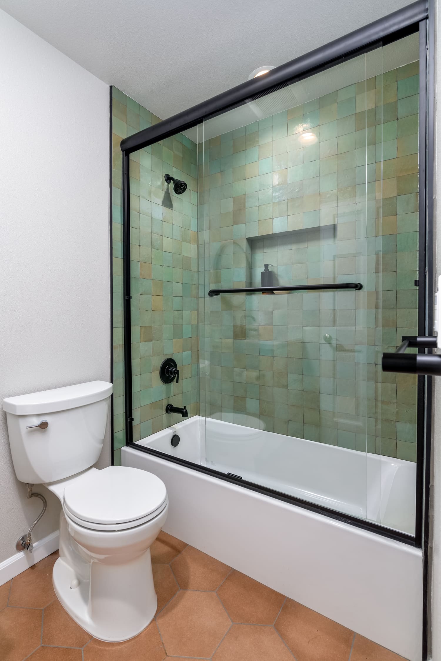 Small bathroom with toilet, bathtub, and glass shower enclosure with green tile.