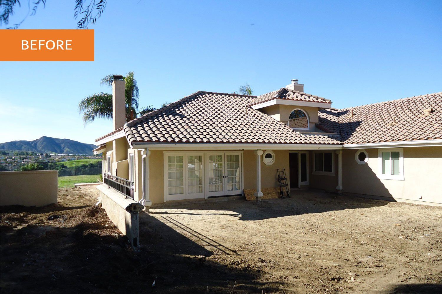 Before photo of a single-story house with a tiled roof and bare yard under a blue sky