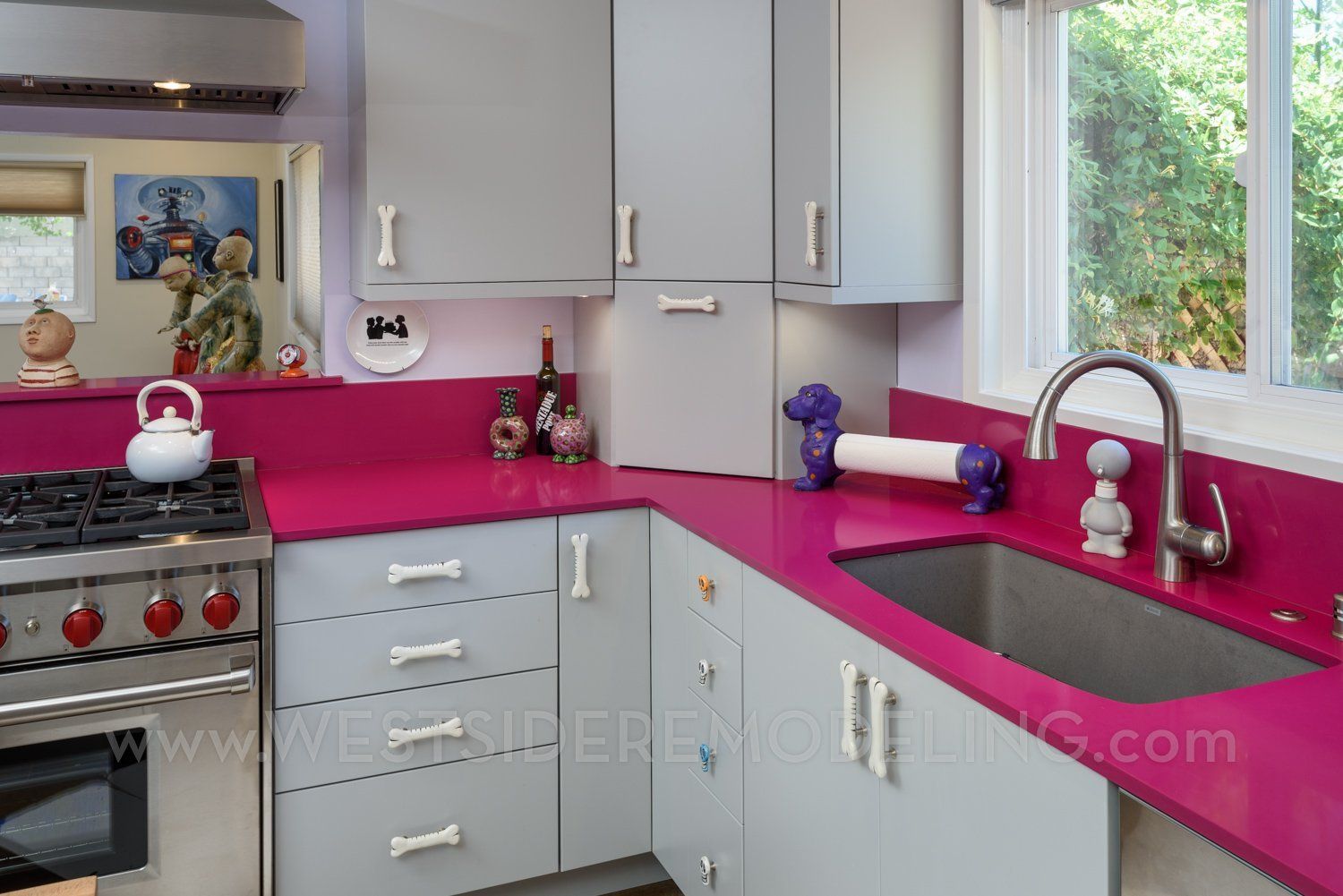 Bright kitchen with gray cabinets, hot pink countertops, stainless stove, and a window over the sink.