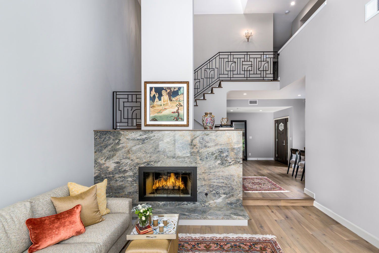Modern living room with stone fireplace, gray sofa, staircase, and bright open hallway