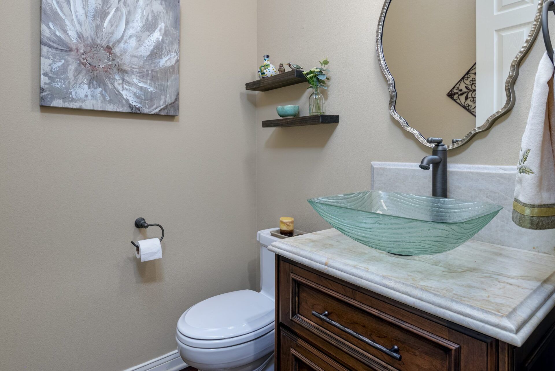 Bathroom vanity with glass sink, mirror, towel ring, and toilet beside a wall shelf and wall art