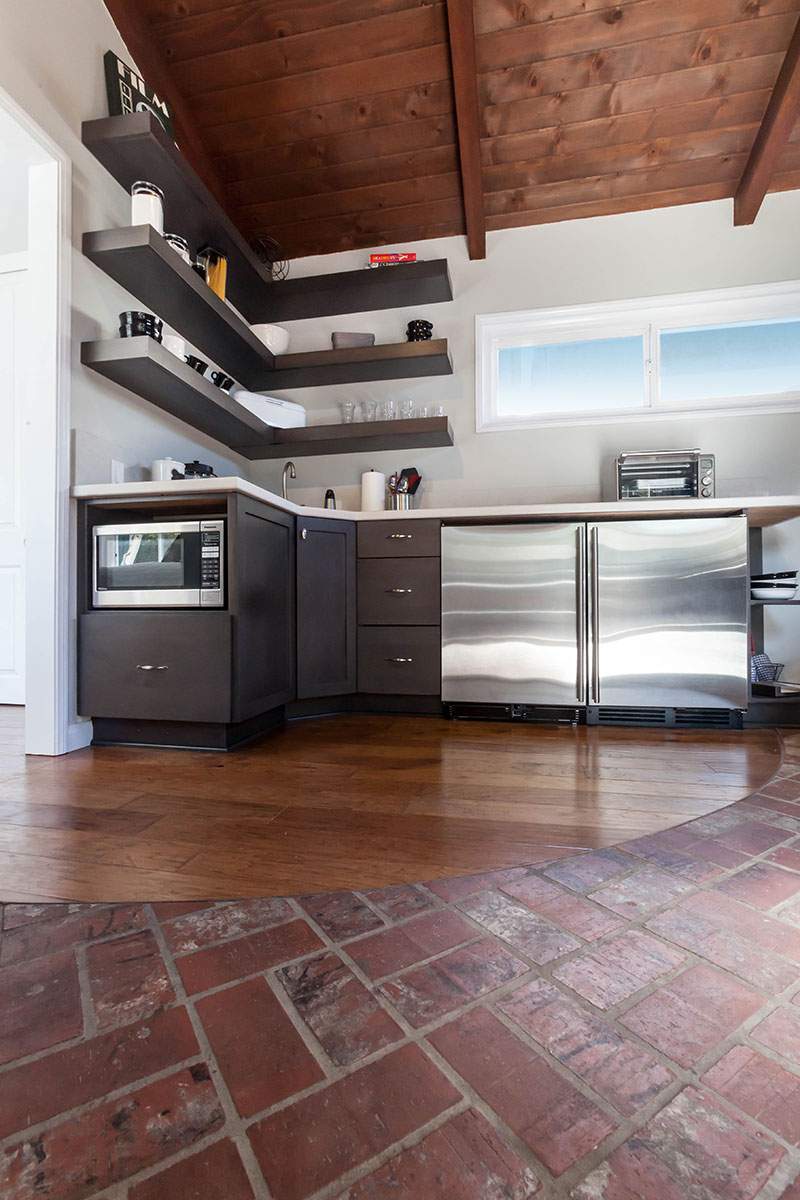 Kitchen with brick floor, stainless steel cabinets, wall shelves, and a stove under a slanted wood ceiling