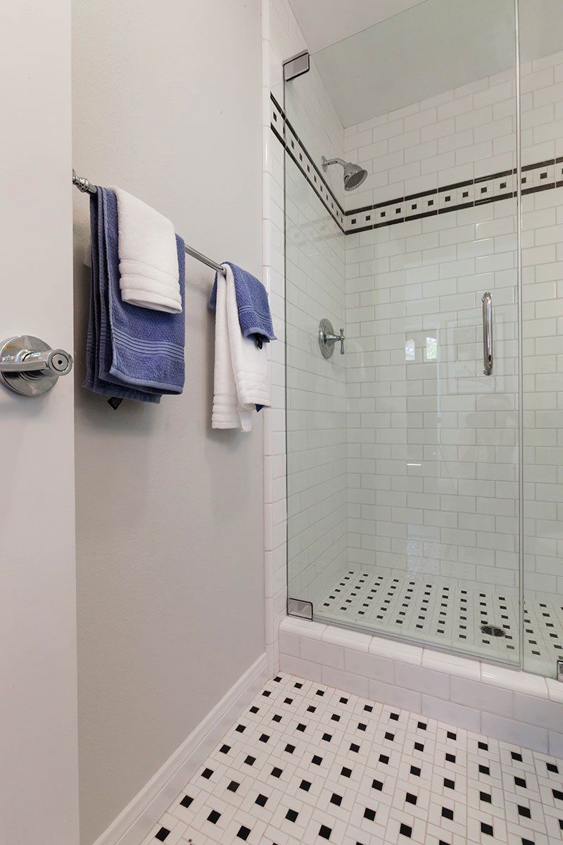 Modern bathroom with glass shower, white tile, and black-and-white patterned floor; towels on wall rack