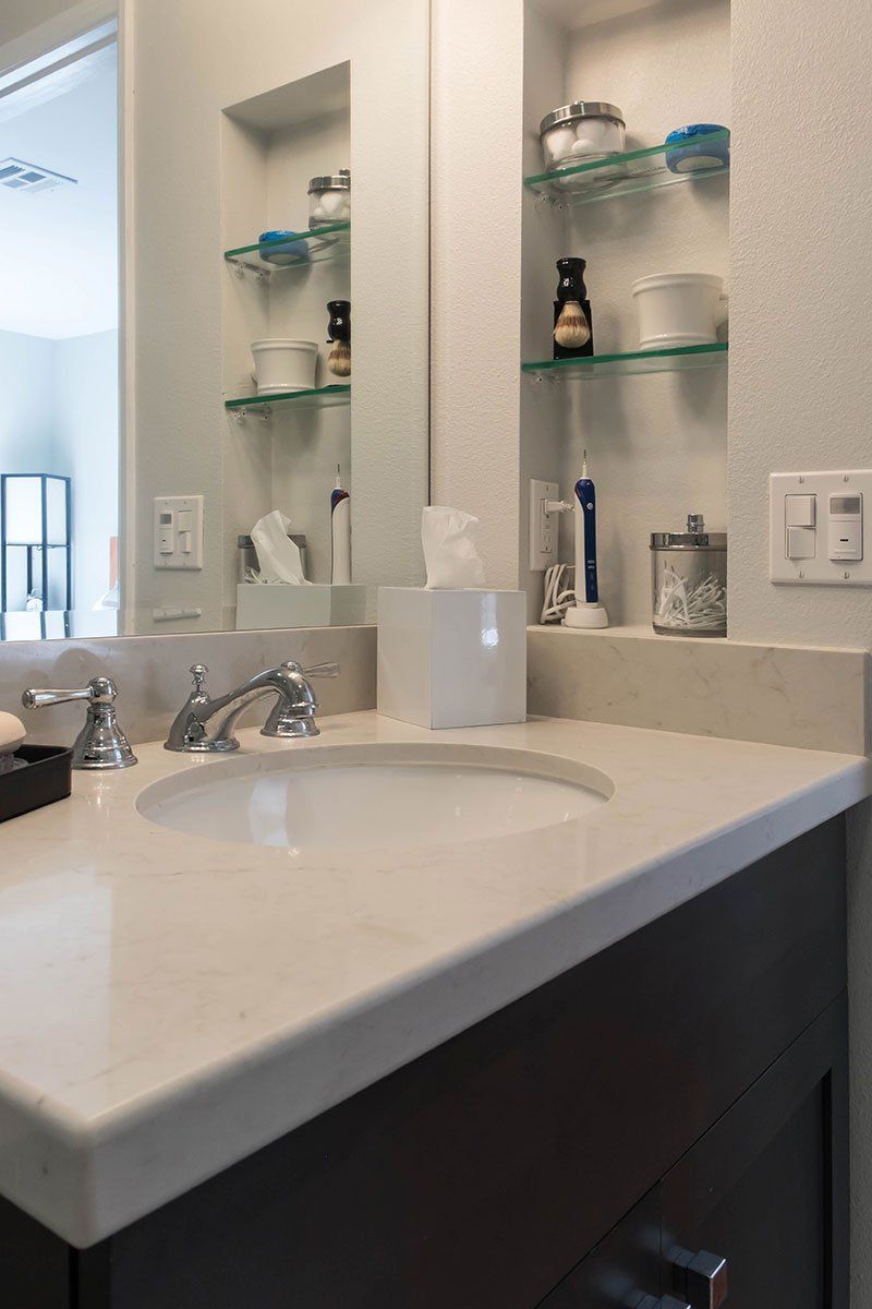 Bathroom sink and mirror with glass shelves, toiletries, and beige tile walls.
