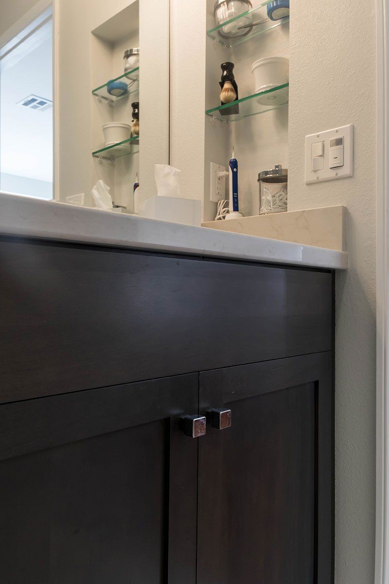 Bathroom vanity with dark cabinet, white countertop, glass shelves, and wall outlet beside a mirror.