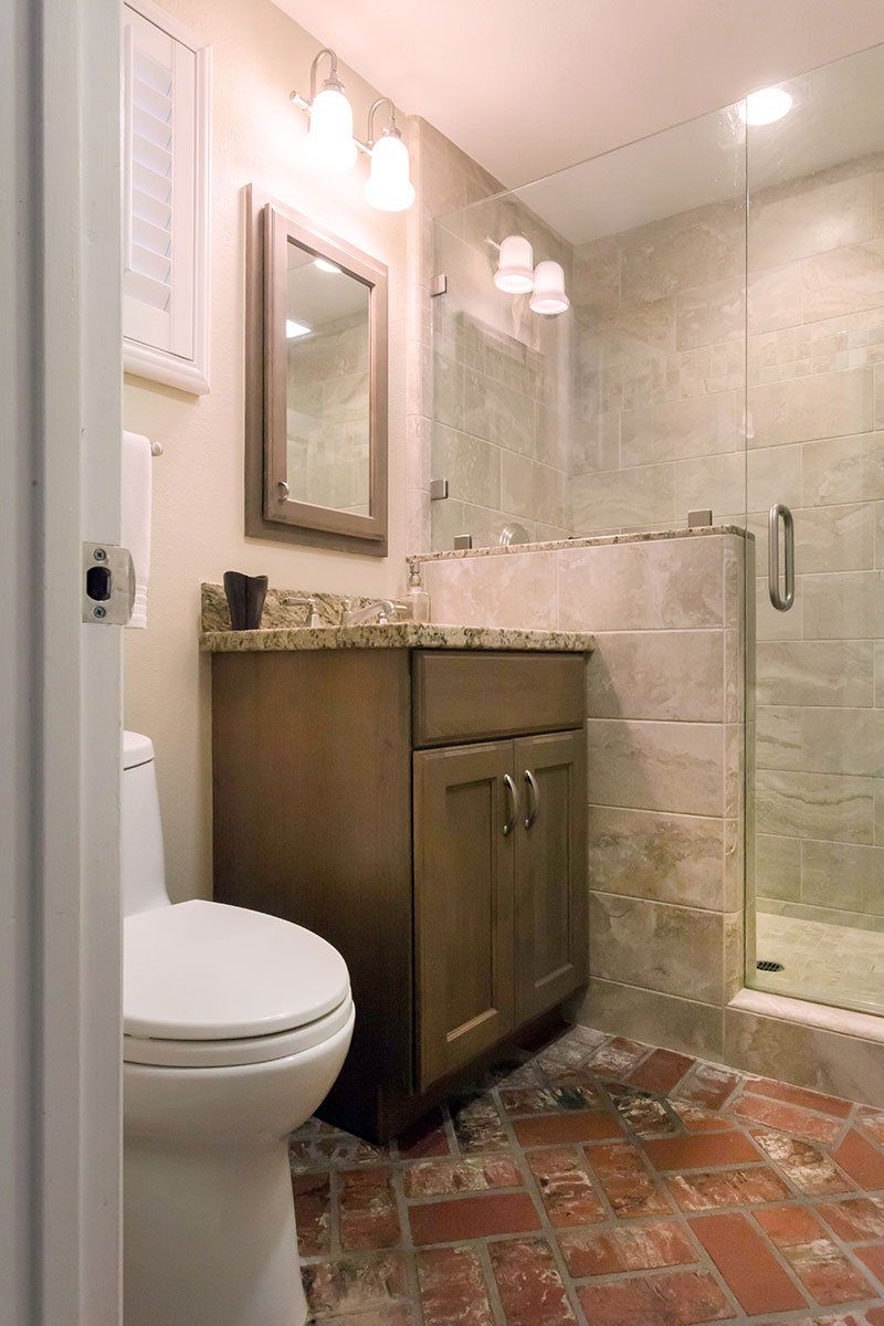 Small bathroom with toilet, vanity, mirror, and glass shower enclosure on terracotta tile floor.