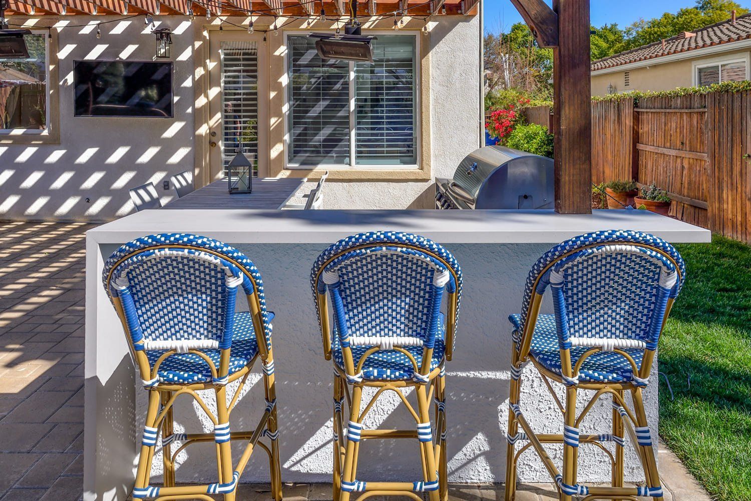 Backyard patio with three blue chairs at a white bar under a pergola, next to a grill and wooden fence.