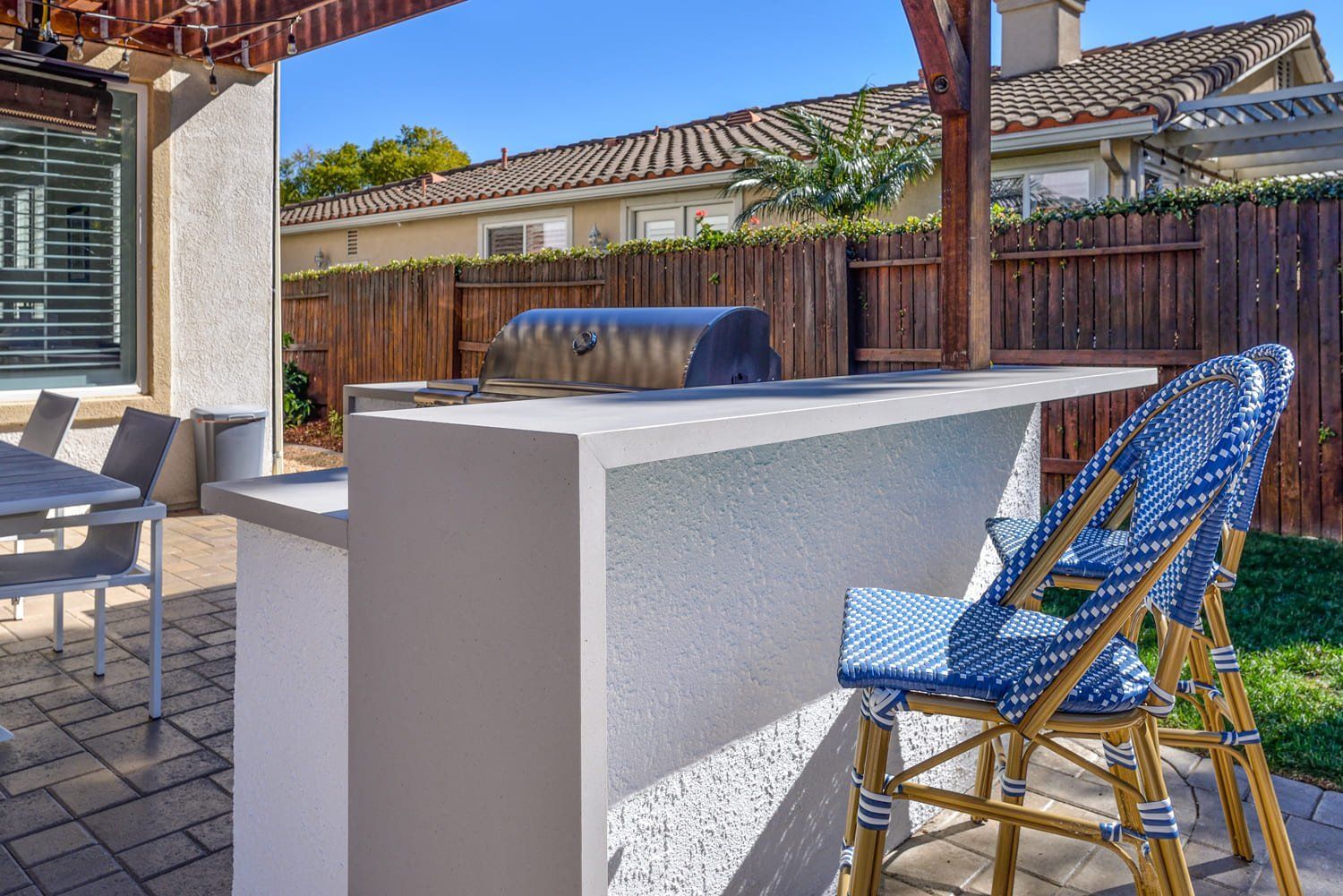 Outdoor patio with a white grill island, blue chair, and covered seating area in a backyard.