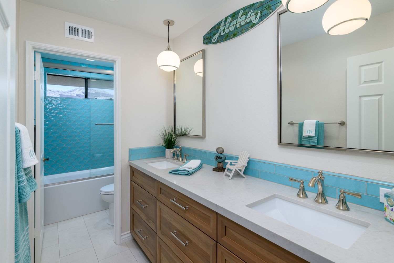 Bright bathroom vanity with wood cabinets, blue accents, large mirror, and shower visible in the background