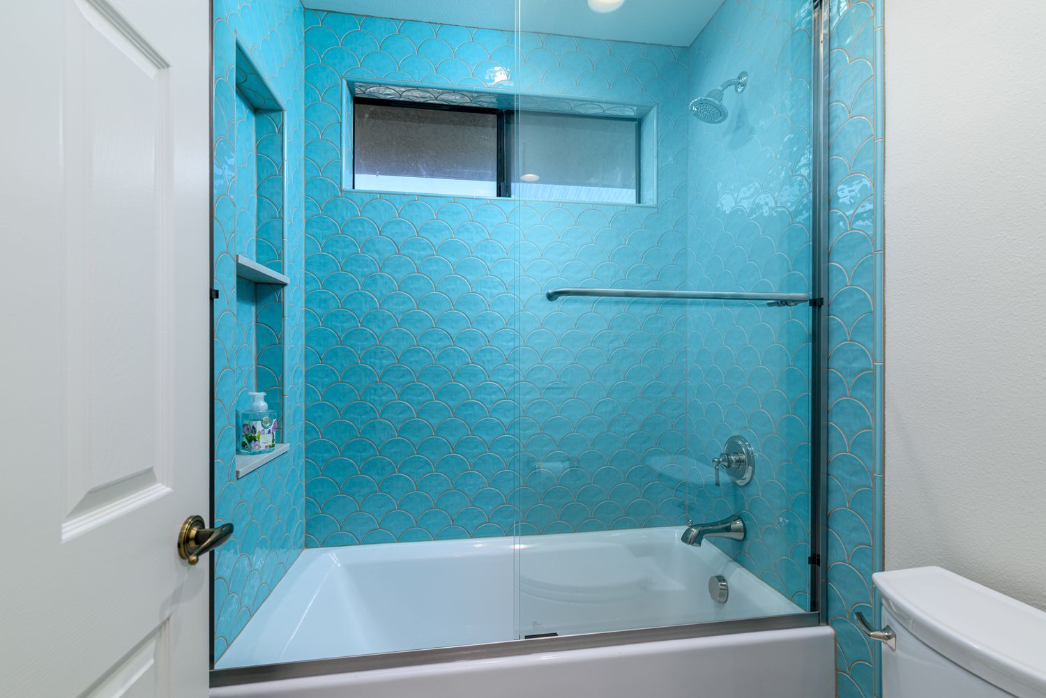 Blue-tiled bathroom shower with bathtub, glass enclosure, and window above the tub