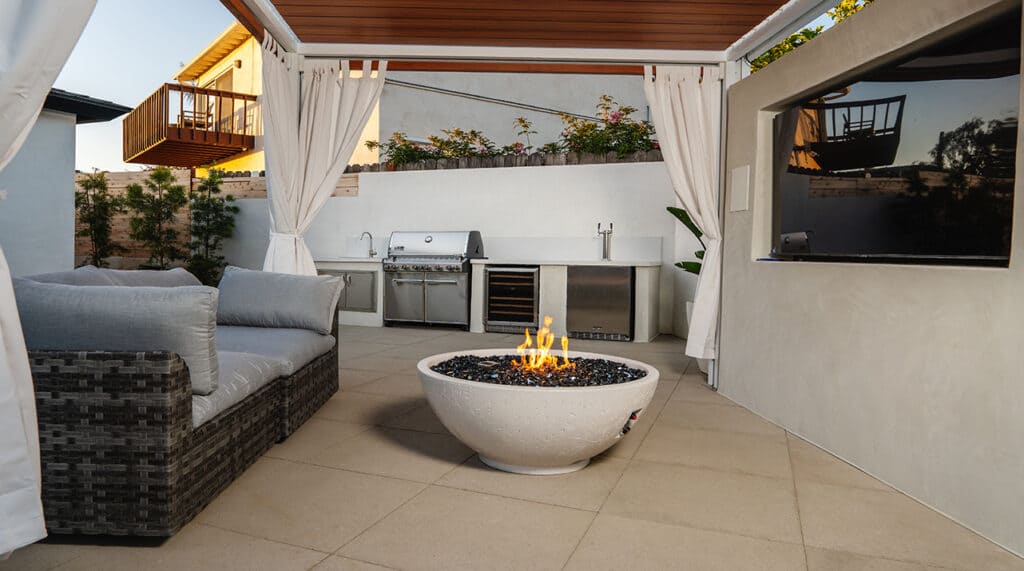 Modern patio with wicker seating, pergola, wall-mounted TV, and a round fire pit at center.