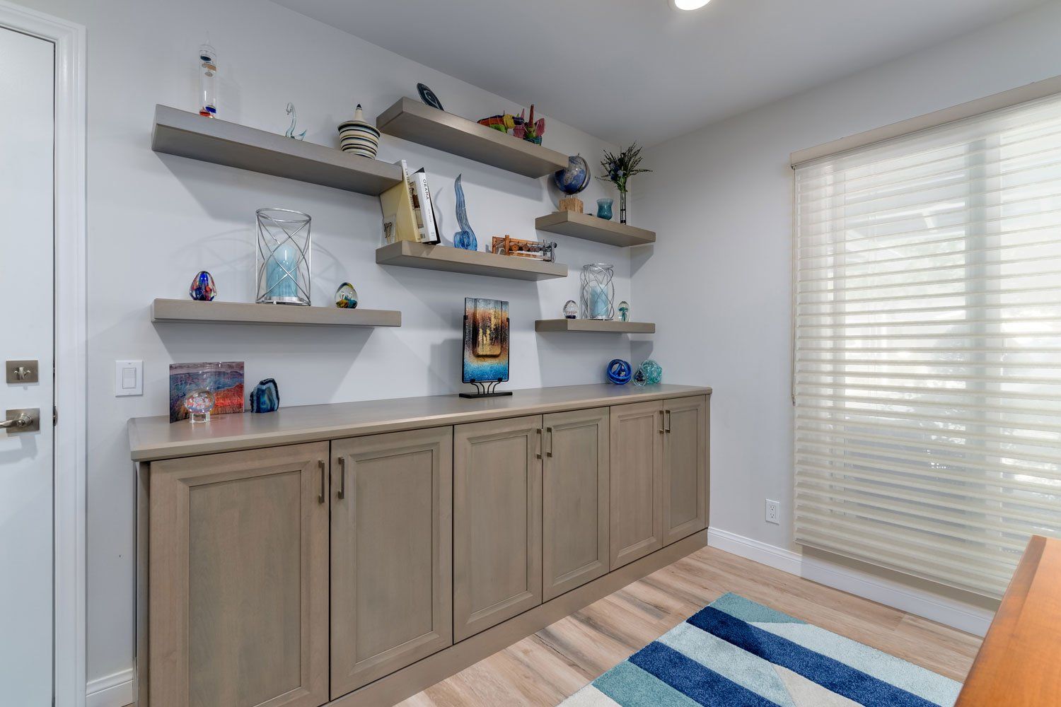 Modern home office with gray cabinets, floating shelves, blue accents, and a striped rug by window blinds