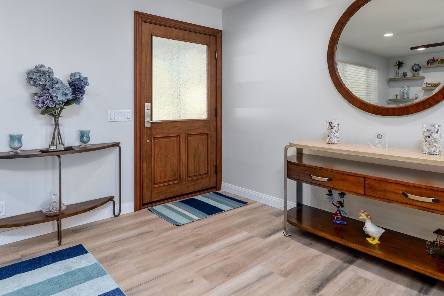 Bright entryway with wooden door, round mirror, console table, and blue rugs on light wood flooring