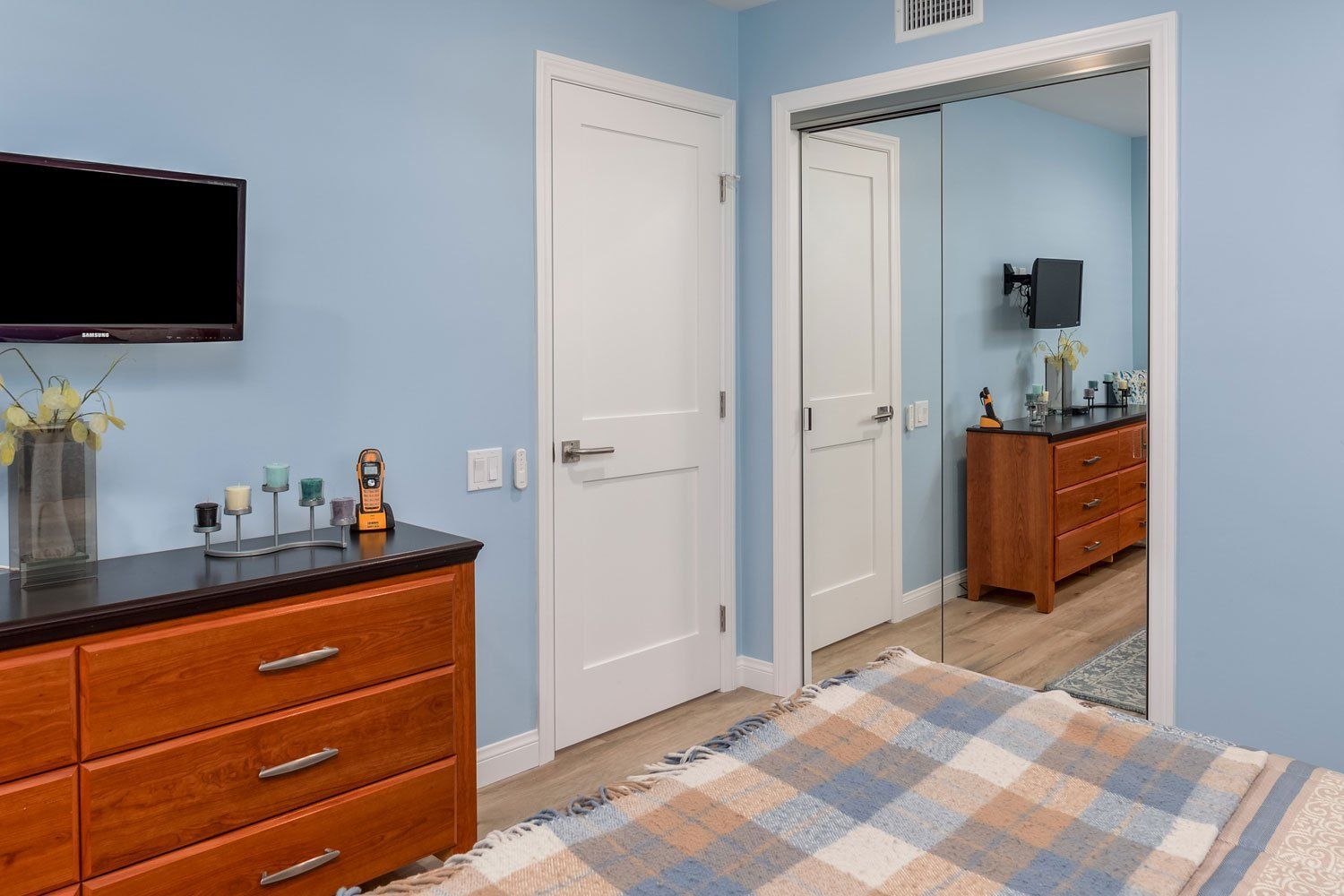 Blue bedroom with dresser, wall-mounted TV, mirrored closet door, and bed edge in foreground