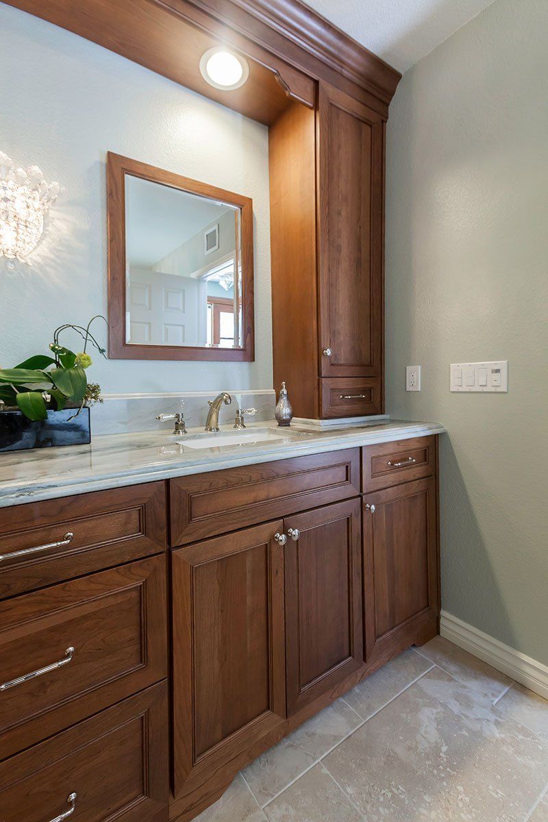 Wooden bathroom vanity with marble countertop, mirror, sink, and wall sconce in a tiled room