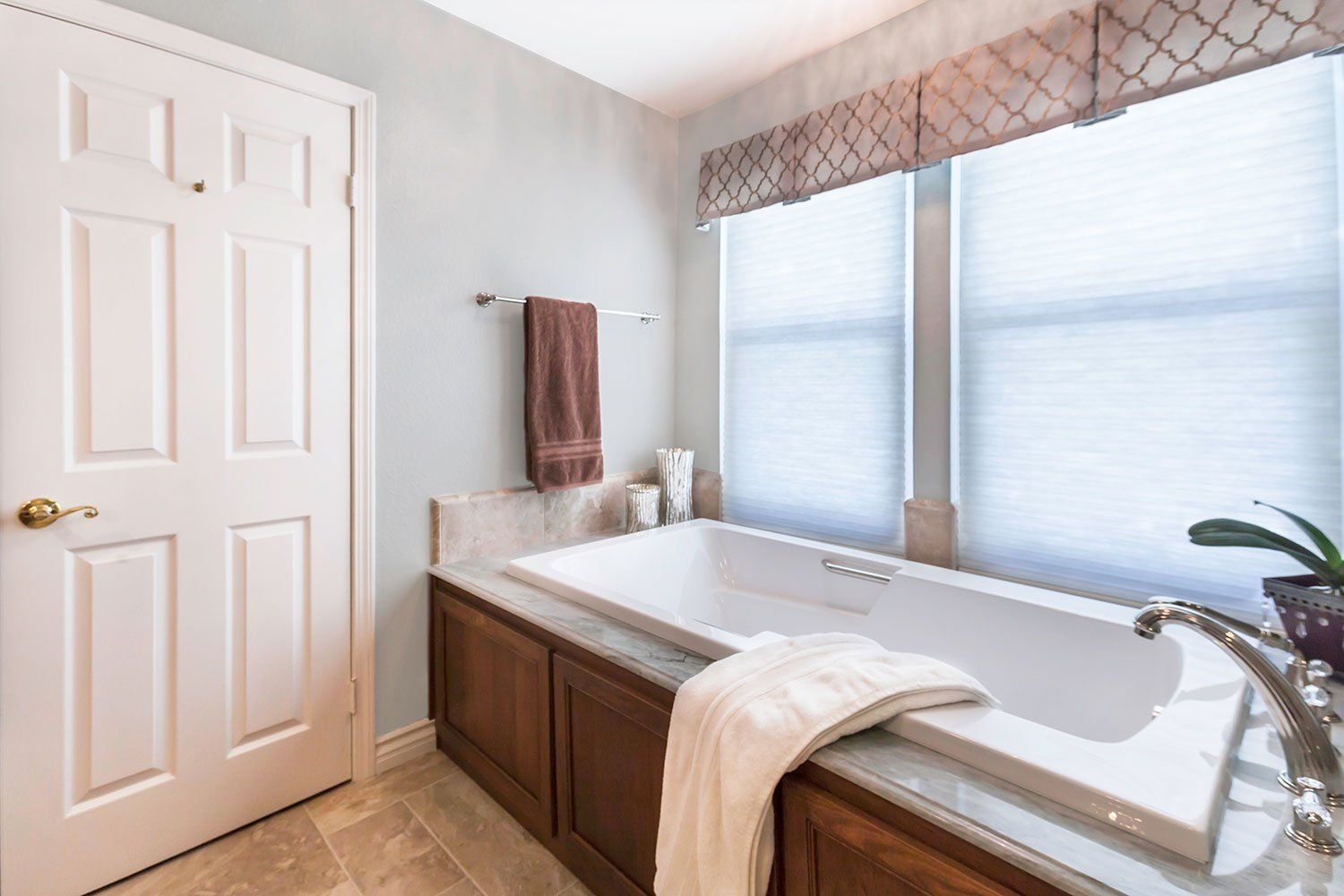 Bright bathroom with a large bathtub, window blinds, brown cabinets, and beige tile floor.