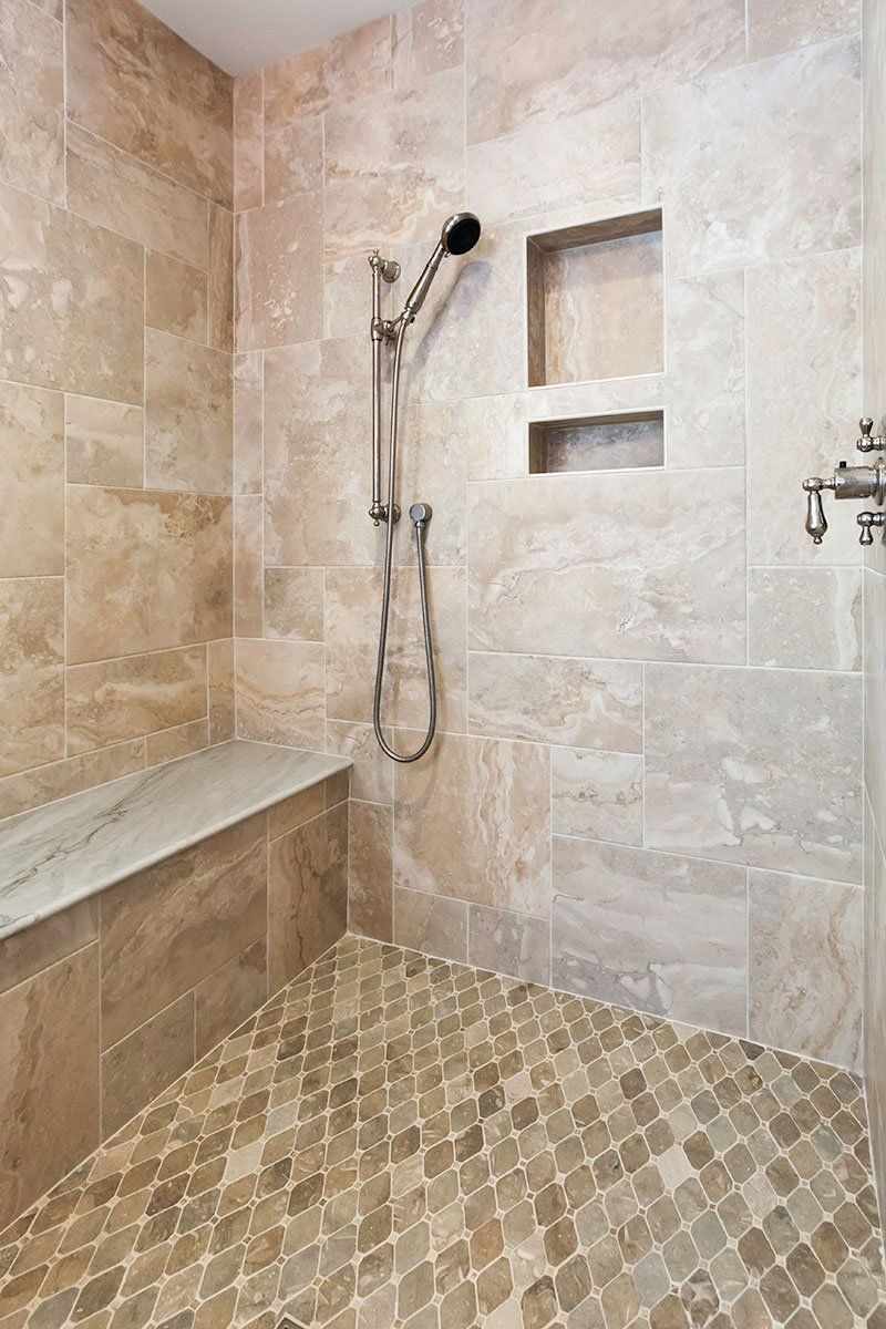 Beige tiled walk-in shower with bench, handheld showerhead, and recessed wall shelf.