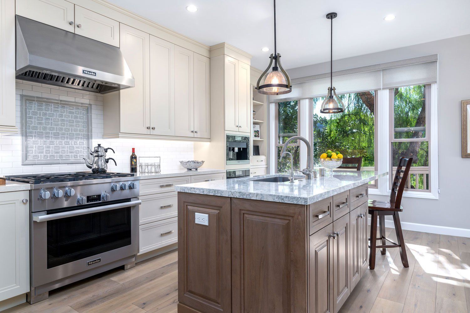 Bright modern kitchen with white cabinets, marble island, stainless stove, and large windows by a breakfast nook