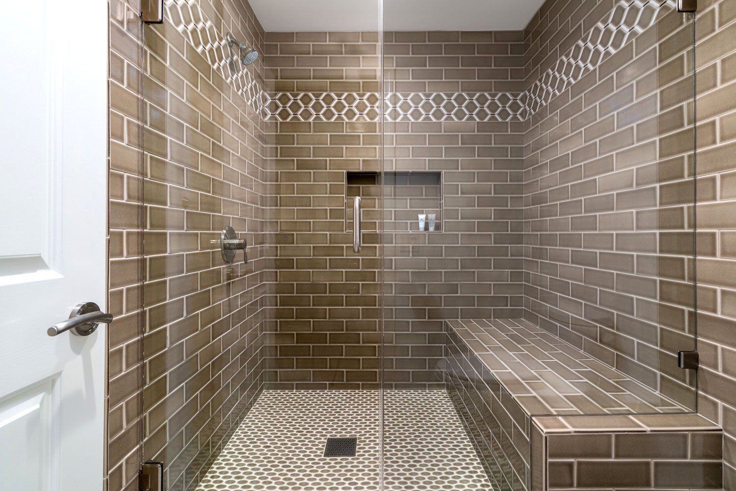 Modern tiled shower with glass enclosure and built-in bench, viewed through an open white door