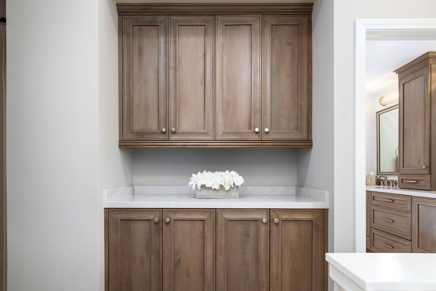 Wooden bathroom vanity with upper cabinets, white countertop, and white flowers in a recessed alcove.