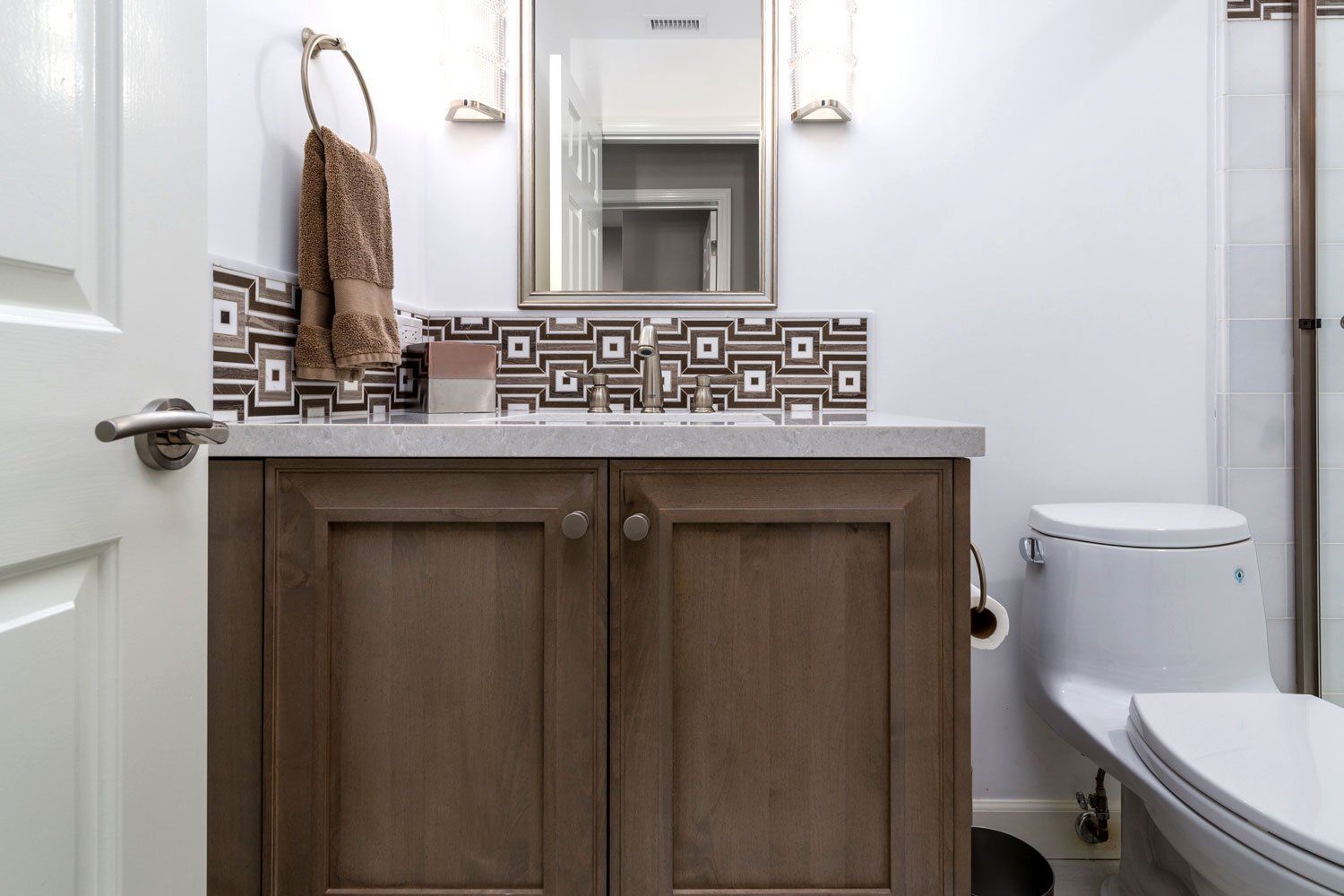 Small bathroom with wood vanity, patterned tile backsplash, mirror, and toilet beside a shower.