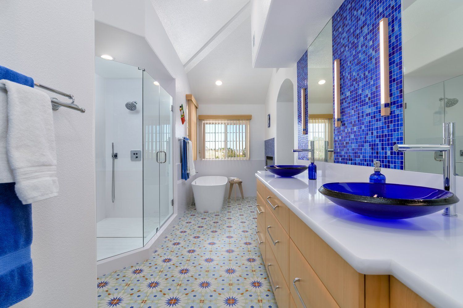 Modern bathroom with blue mosaic tiles, double sink vanity, glass shower, and patterned floor tiles