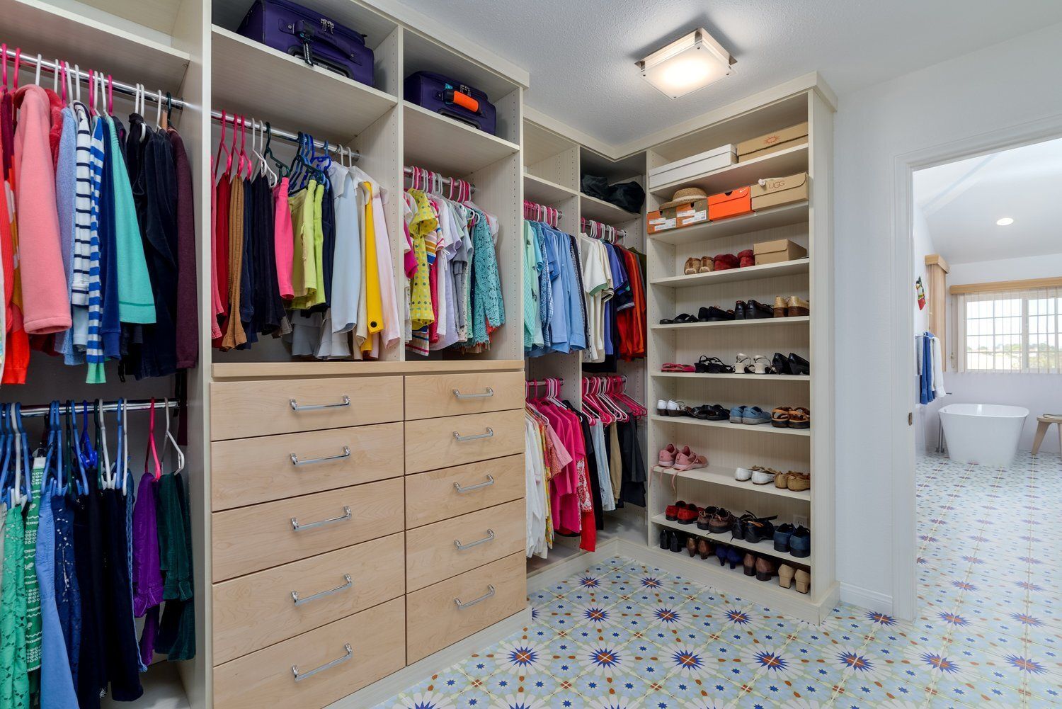 Bright walk-in closet with colorful hanging clothes, drawers, shoe shelves, and a doorway to a bathroom.