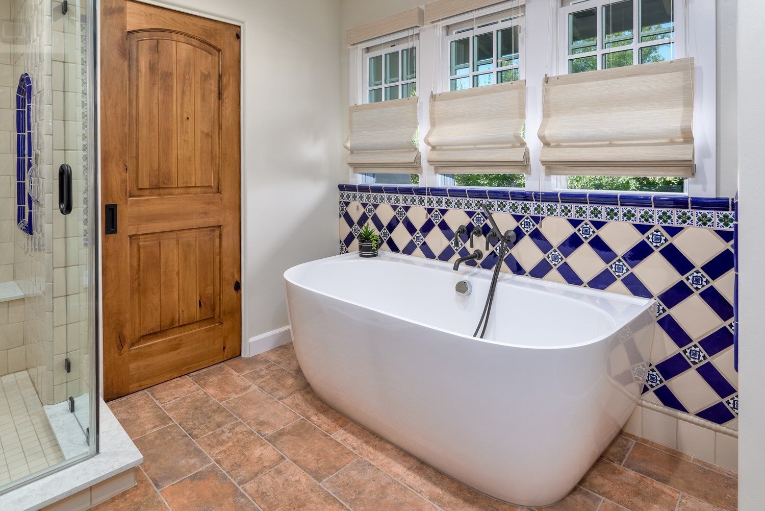 Modern bathroom with freestanding tub, blue-and-white tile wall, wooden door, and window blinds