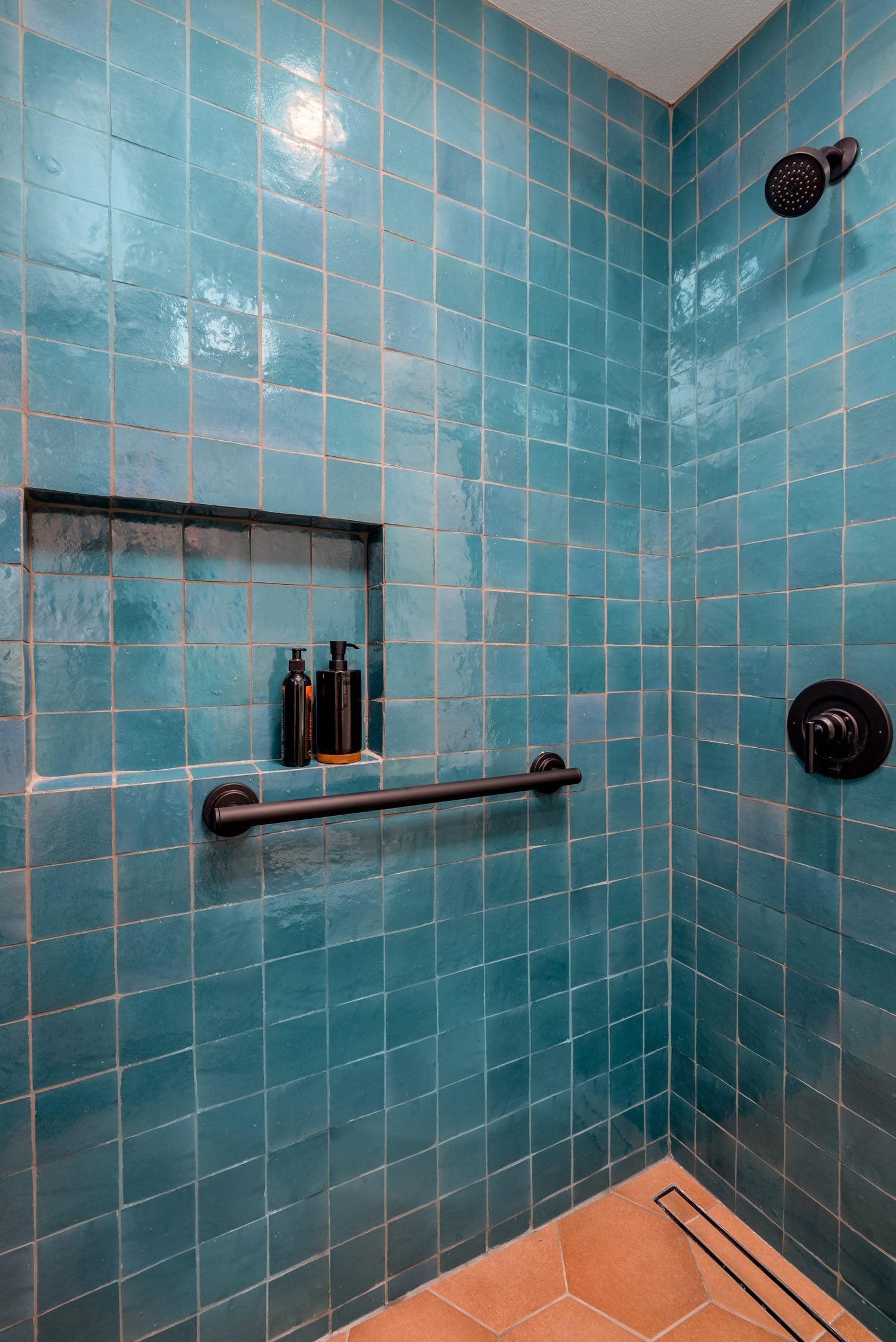 Blue-tiled shower stall with built-in shelf, grab bar, and tan floor tiles