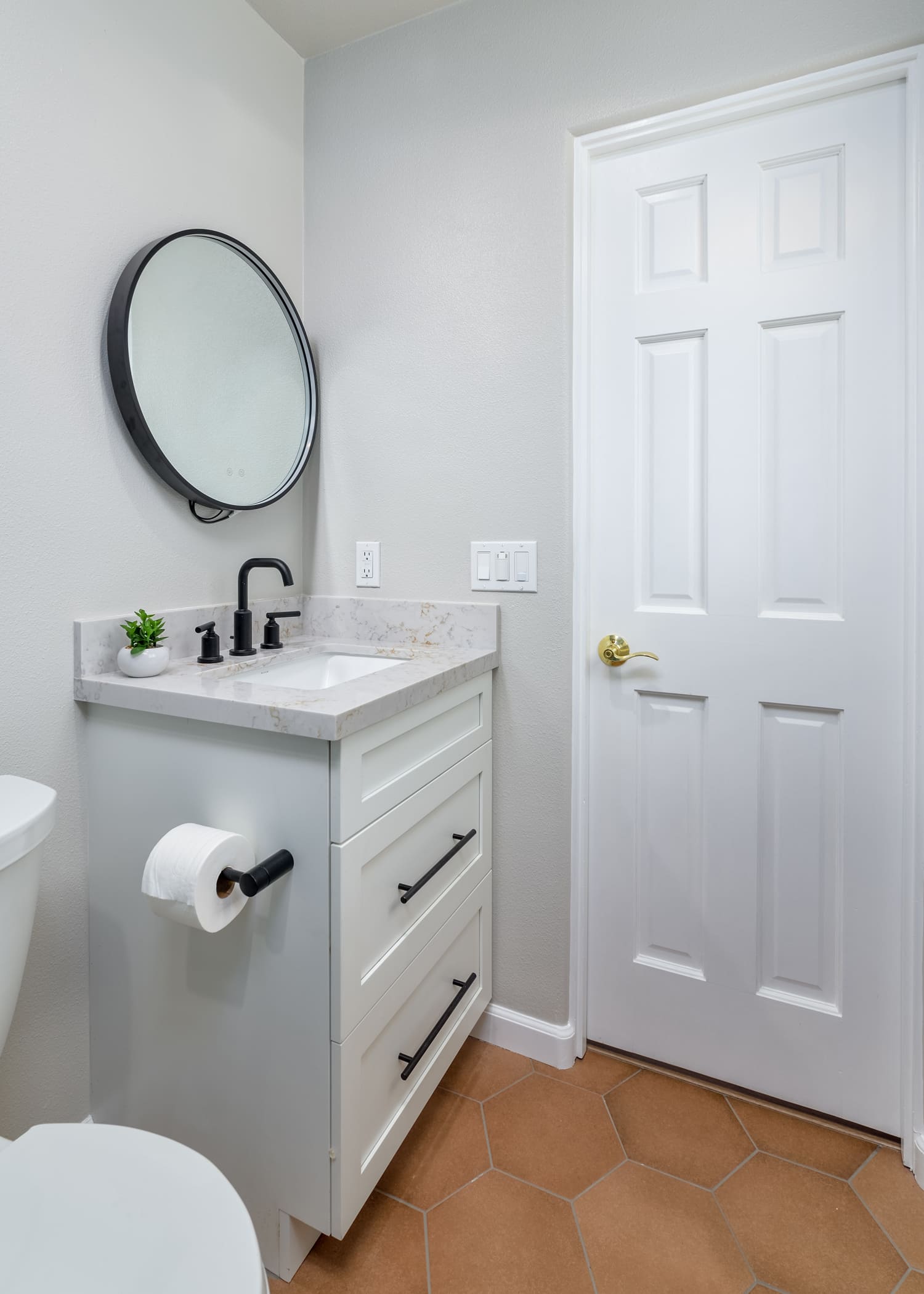 Small white bathroom with vanity sink, round mirror, toilet, and closed door on tan tile floor
