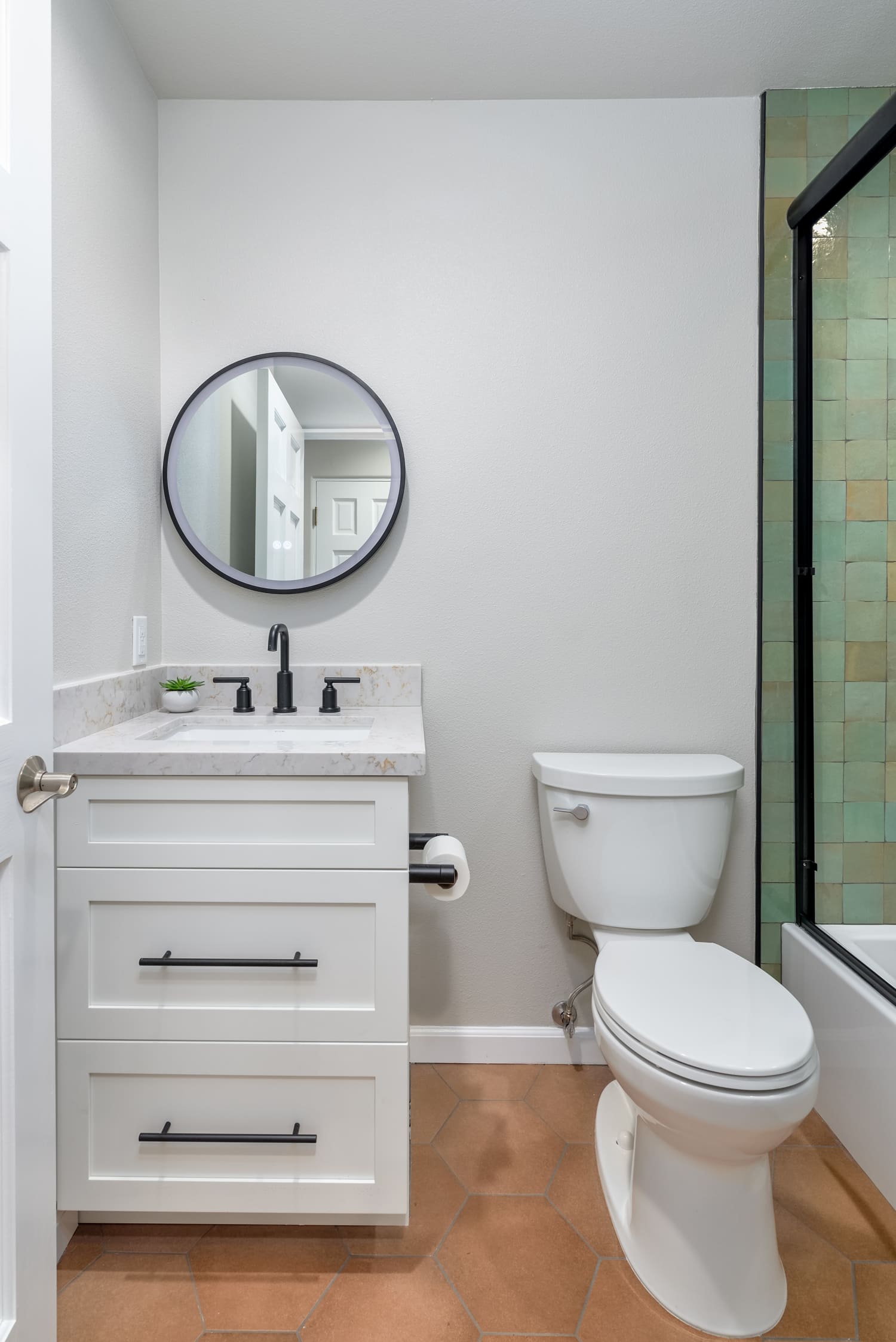 Small bathroom with white vanity, round mirror, toilet, and glass shower door; light wood floor.