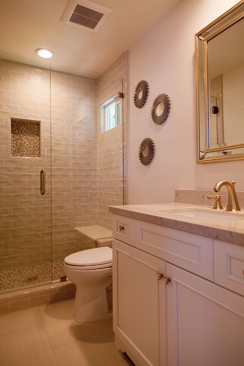 Small beige bathroom with glass shower, toilet, vanity, and gold-framed mirror