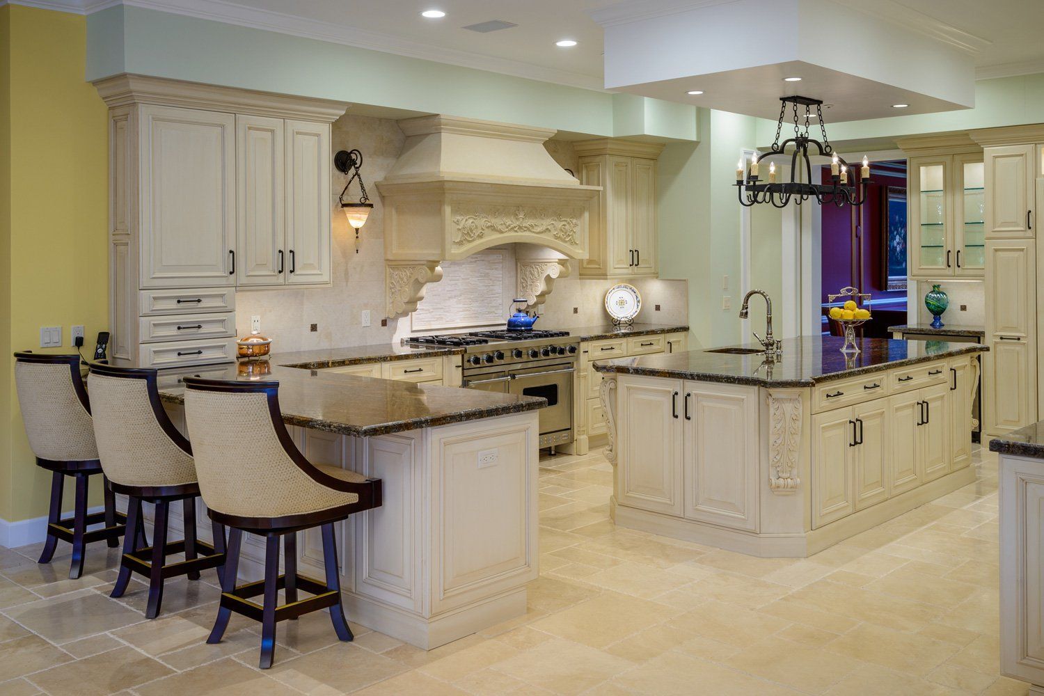 Elegant cream-colored kitchen with granite island, bar stools, and stainless hood under recessed lighting